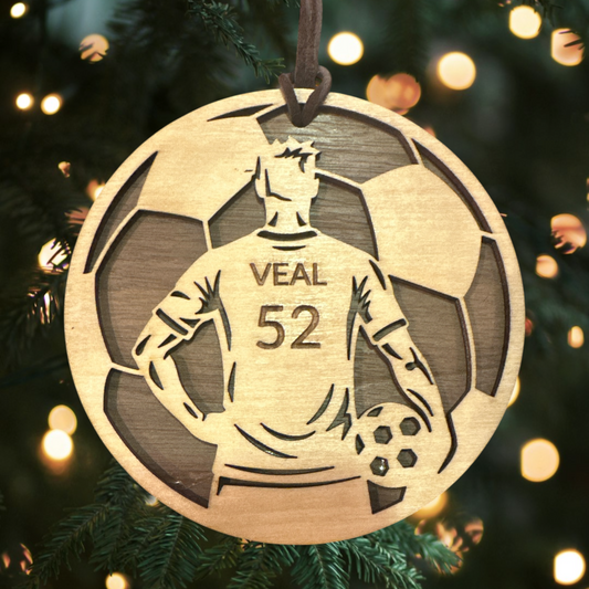Wooden ornament with a soccer player design and 'VEAL 52' text, hanging on a Christmas tree.